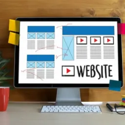 Monitor wyświetla szkic strony internetowej z notatkami, obok kubek, roślina doniczkowa i okulary na biurku.