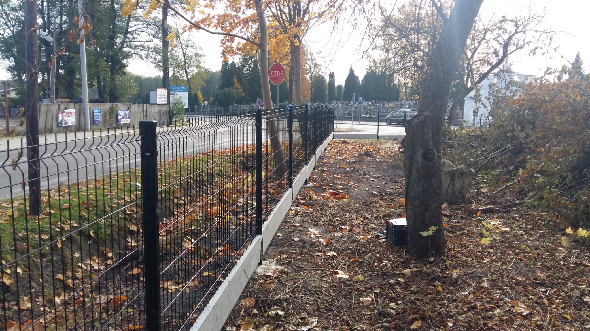 Nowo zainstalowane czarne ogrodzenie panelowe z betonową podmurówką, oddzielające drogę od terenu z opadłymi liśćmi, widoczny znak STOP i fragment cmentarza w tle.