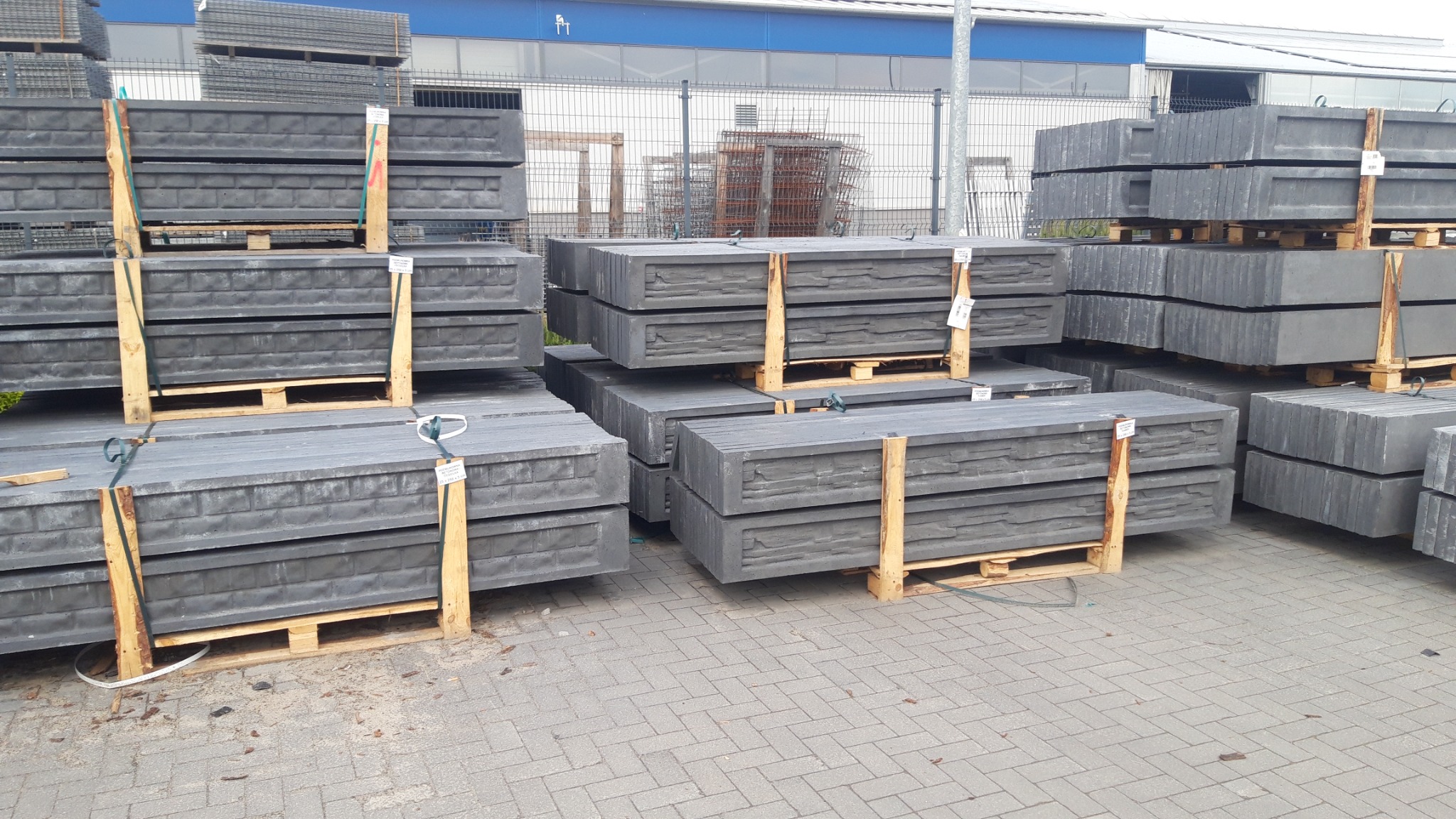 Szare, betonowe elementy ogrodzeniowe imitujące kamień, ułożone na drewnianych paletach, przygotowane do transportu, na tle szarej kostki brukowej i hali magazynowej.
