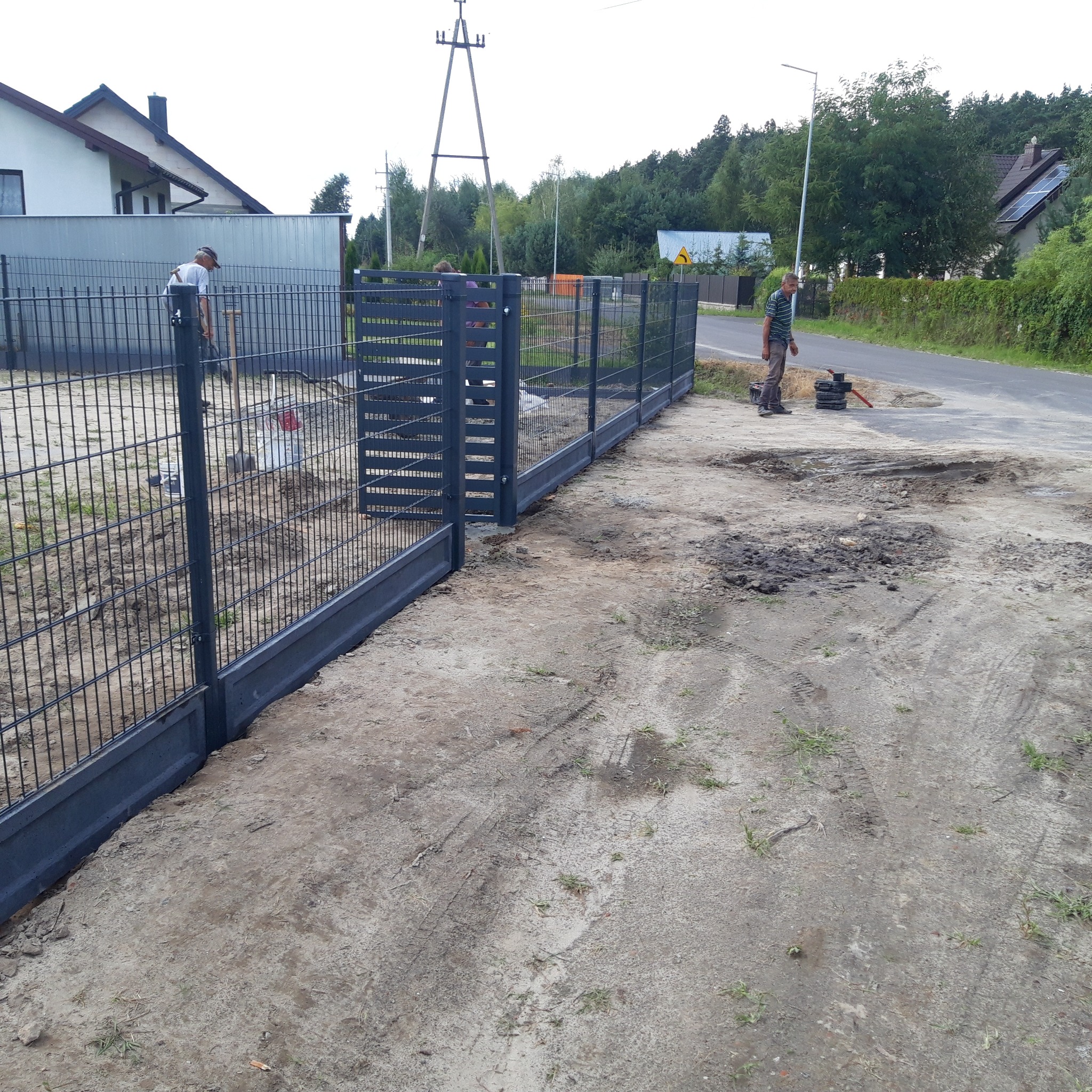 Montaż nowoczesnego, antracytowego ogrodzenia panelowego z podmurówką prefabrykowaną; widoczni pracownicy i narzędzia na placu budowy.