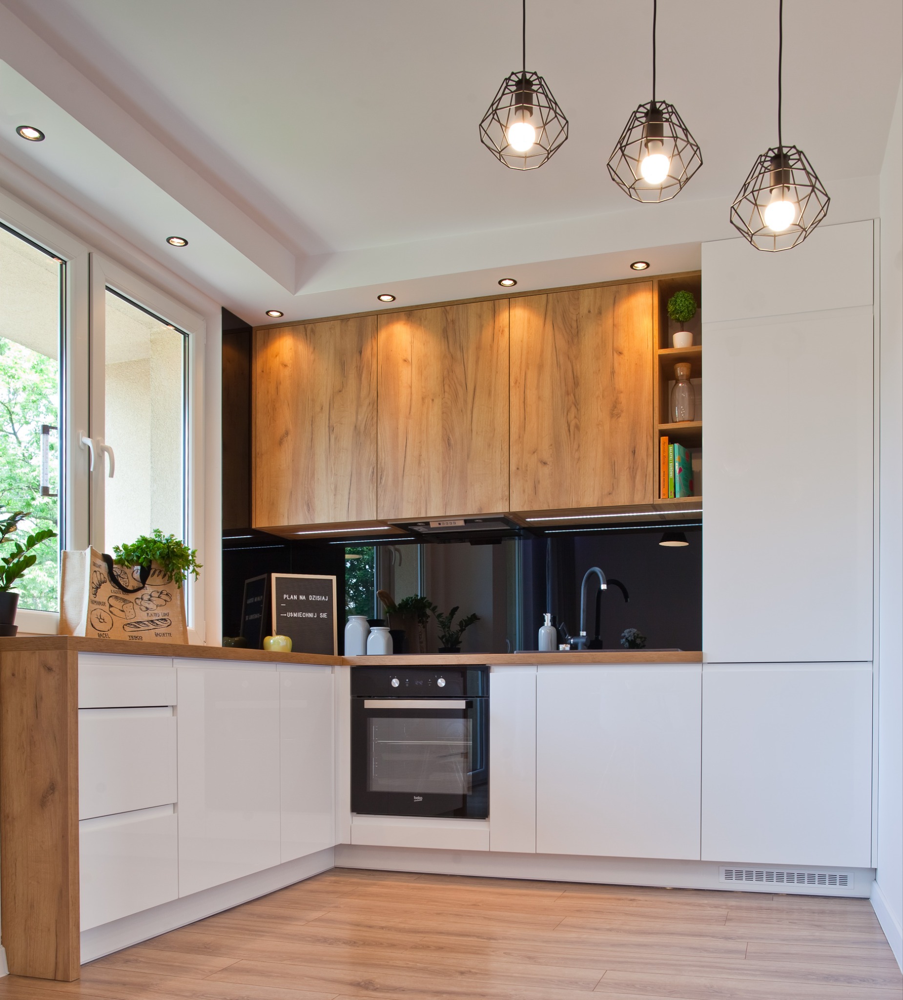 Nowoczesna kuchnia z białymi lakierowanymi frontami i drewnianymi szafkami górnymi, czarnym panelem nad blatem, oświetleniem punktowym i trzema lampami wiszącymi w geometrycznych kloszach.