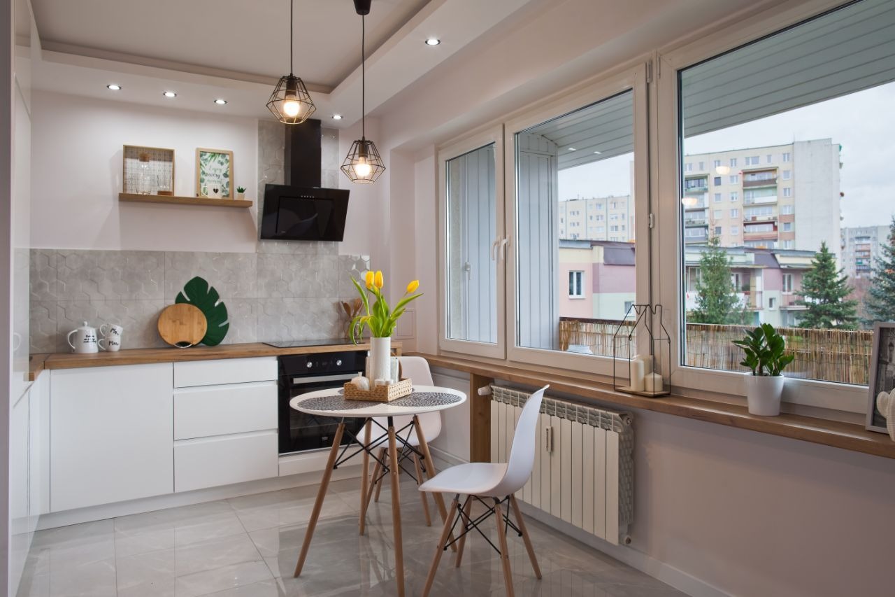 Jasna kuchnia z białymi szafkami, drewnianym blatem i okrągłym stołem z tulipanami, widok na osiedle mieszkaniowe za oknem.