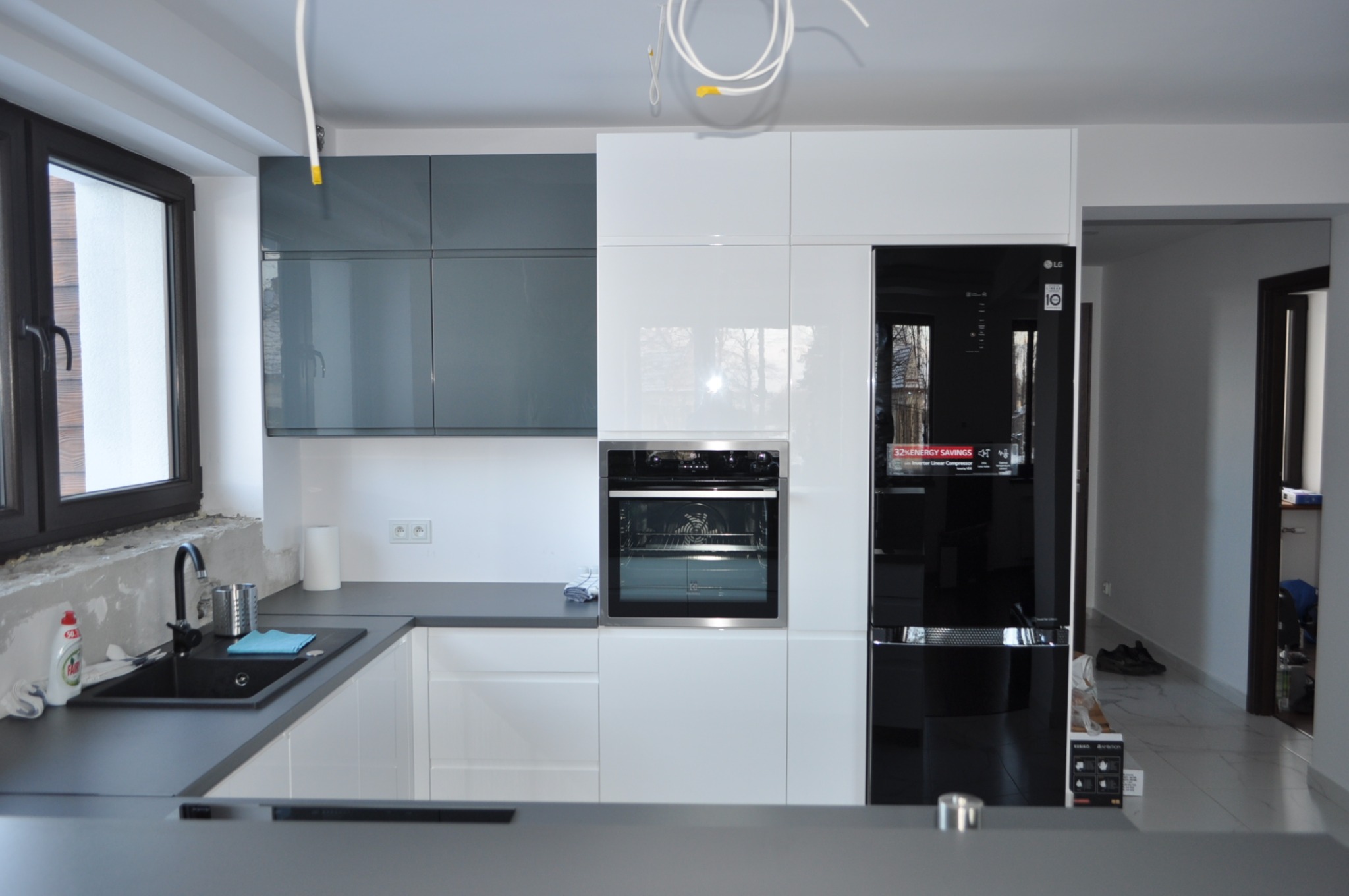 Nowoczesna kuchnia z szarymi i białymi szafkami, czarnym zlewem i lodówką LG, częściowo wykończona ściana nad zlewem, widoczne kable na suficie.