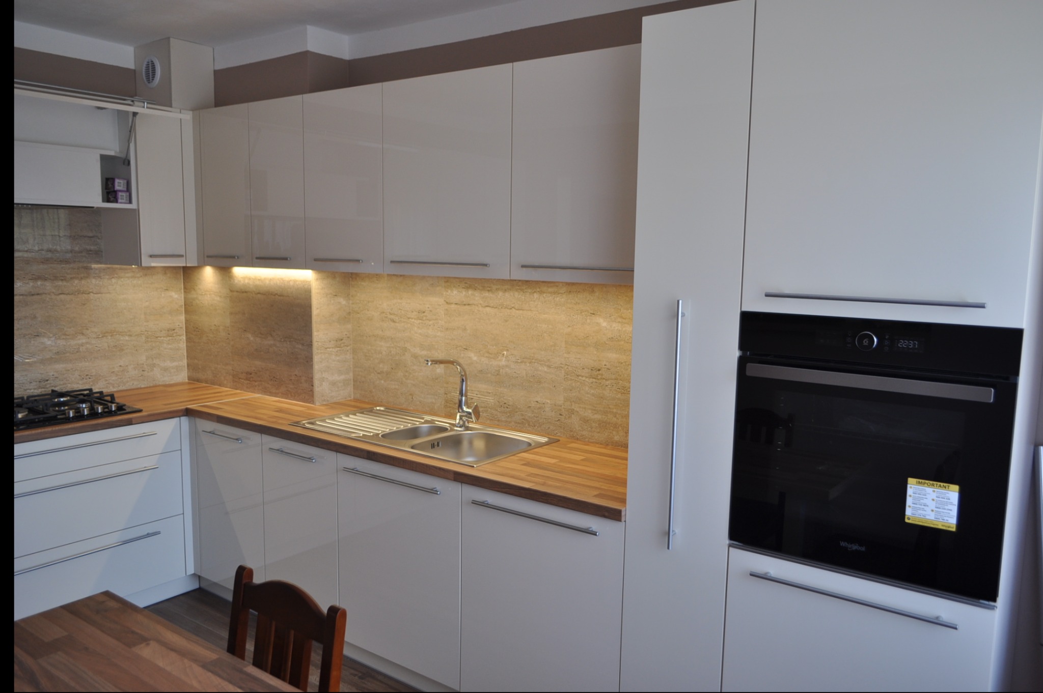 Nowoczesna, jasna kuchnia z błyszczącymi frontami, drewnianym blatem i marmurową ścianą nad blatem, z piekarnikiem w zabudowie.