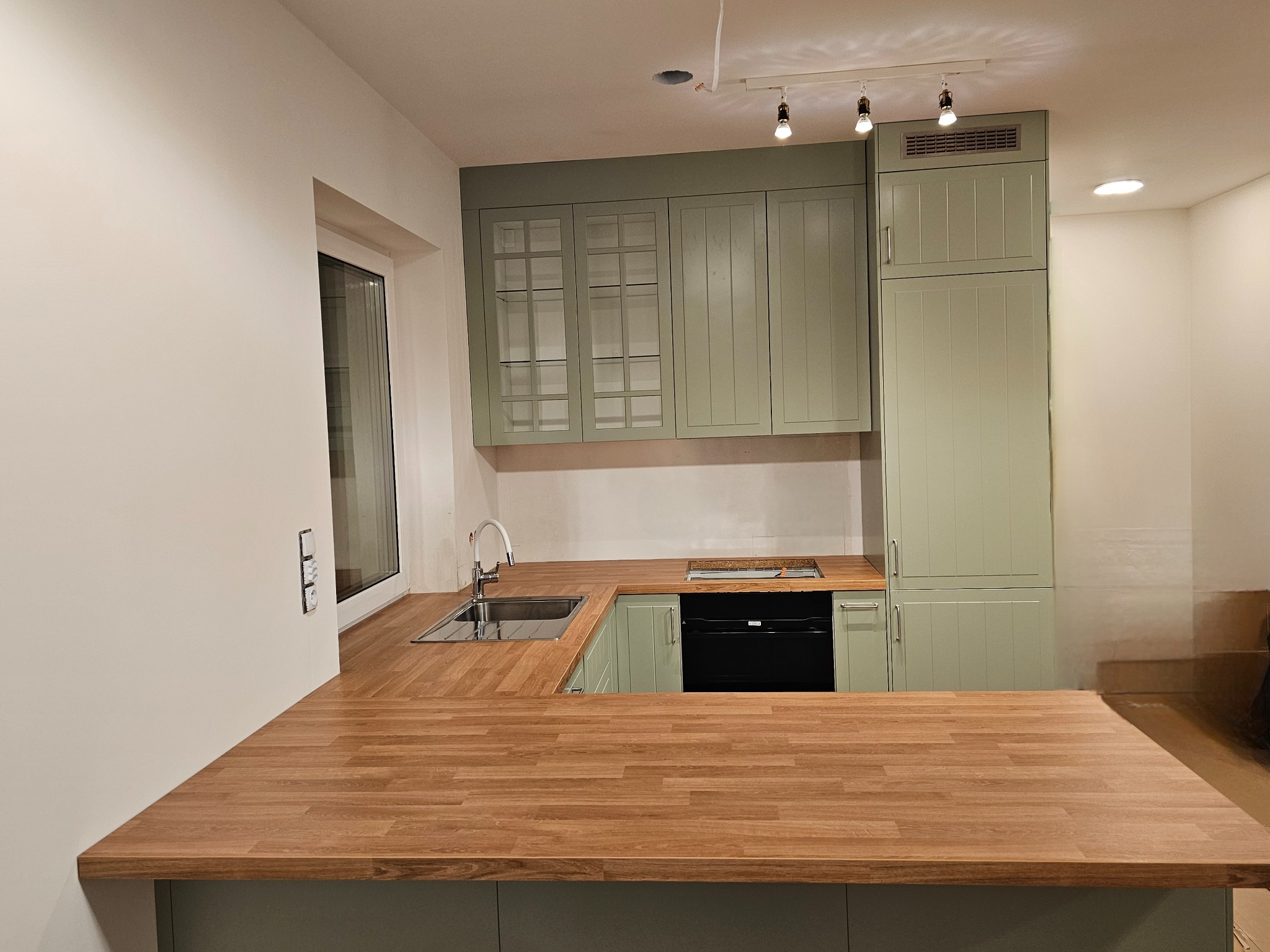 Nowoczesna kuchnia w odcieniach zieleni z drewnianym blatem, widok na zlewozmywak, płytę grzewczą i oświetlenie punktowe.