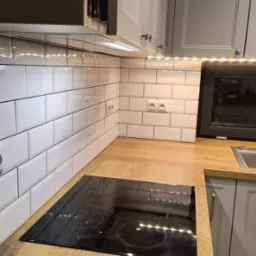 Kuchnia po remoncie: białe, prostokątne płytki na ścianie nad blatem z drewnianym dekorem, widoczne gniazdka elektryczne i płyta indukcyjna, szare szafki z chromowanymi uchwytami, oświetlenie LED...