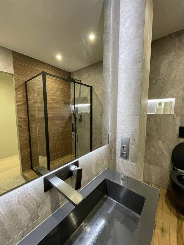 Nowoczesna łazienka z szarą, prostokątną umywalką, baterią montowaną na lustrze, kabiną prysznicową z czarnymi ramami i płytkami imitującymi kamień na ścianach.