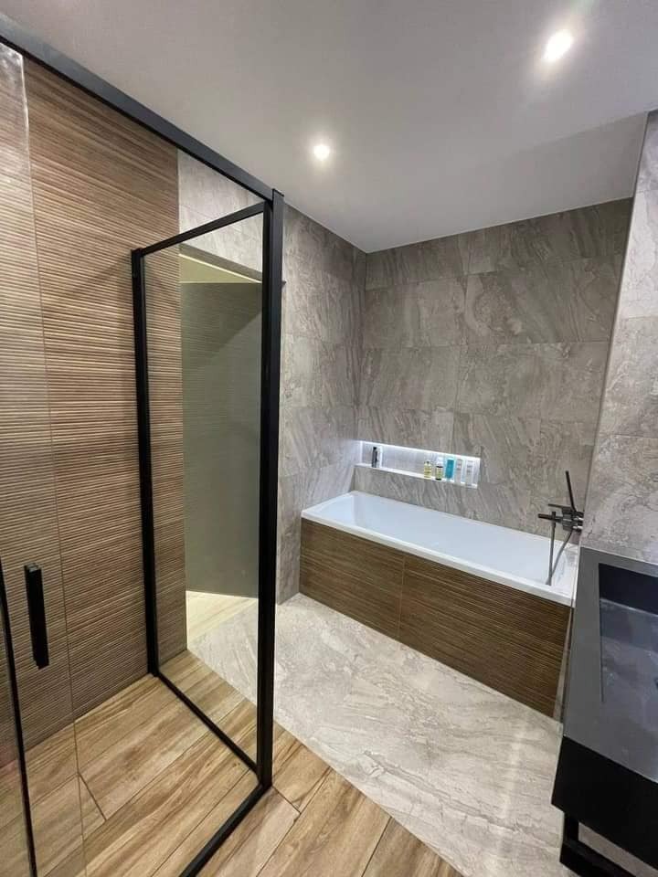 Nowoczesna łazienka z kabiną prysznicową typu walk-in z czarną ramą, wanną obudowaną drewnopodobnymi panelami, szarą marmurową podłogą i wnęką z oświetleniem LED nad wanną.