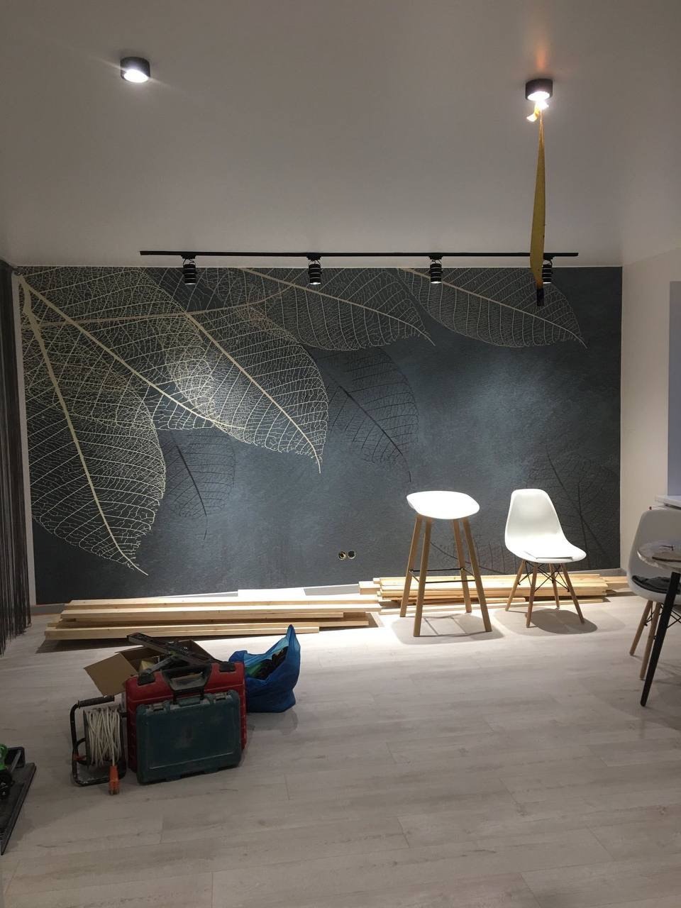 Ściana z szarym deseniem liści, oświetlenie szynowe, dwie białe krzesła i narzędzia na podłodze podczas prac wykończeniowych.