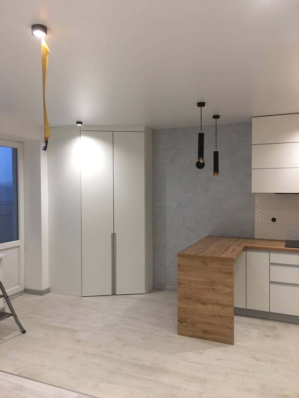 Nowoczesna kuchnia w jasnych barwach z drewnianym blatem, minimalistycznymi lampami wiszącymi i ukrytą szafą w rogu pomieszczenia, widok z poziomu podłogi.