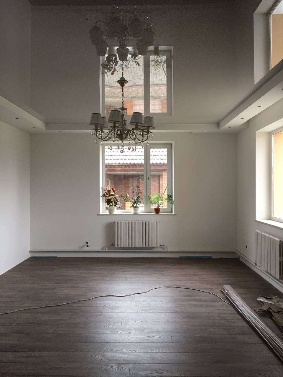 Pusty pokój z ciemną podłogą, białymi ścianami, grzejnikami pod oknami z roślinami, dekoracyjnym żyrandolem i listwami przypodłogowymi w trakcie montażu