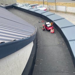 Waterproof Szymon Ptak - Narzędzia dekarskie, w tym wiertarka akumulatorowa i torba narzędziowa Milwaukee, na płaskim, czarnym dachu z obróbkami blacharskimi w kolorze szarym, widok z góry, w tle zabudowa miejska.