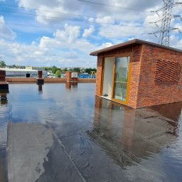 Waterproof Szymon Ptak - Widok na płaski dach z wykończeniem z cegły klinkierowej, z oknem dachowym i elementami wentylacyjnymi. Na dachu widoczne odbicia nieba i chmur.