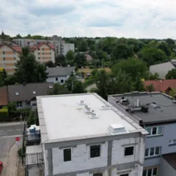Biały dach płaski z kominkami wentylacyjnymi na budynku w stanie surowym otwartym, widok z góry na tle zabudowy miejskiej i zieleni.