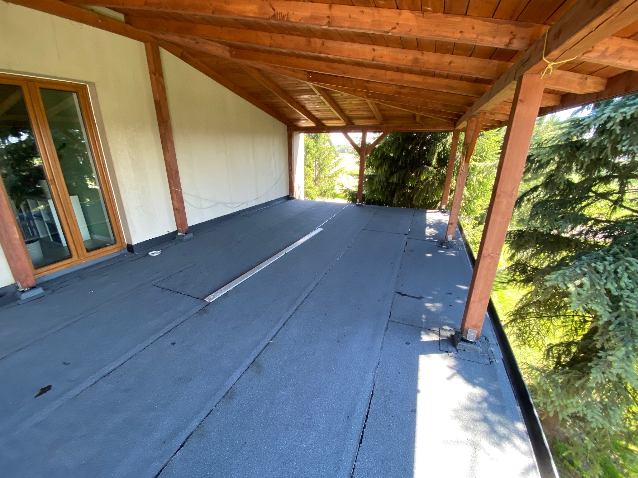 Widok z góry na płaski dach pokryty papą termozgrzewalną, z drewnianą konstrukcją zadaszenia i widokiem na zieleń w tle.