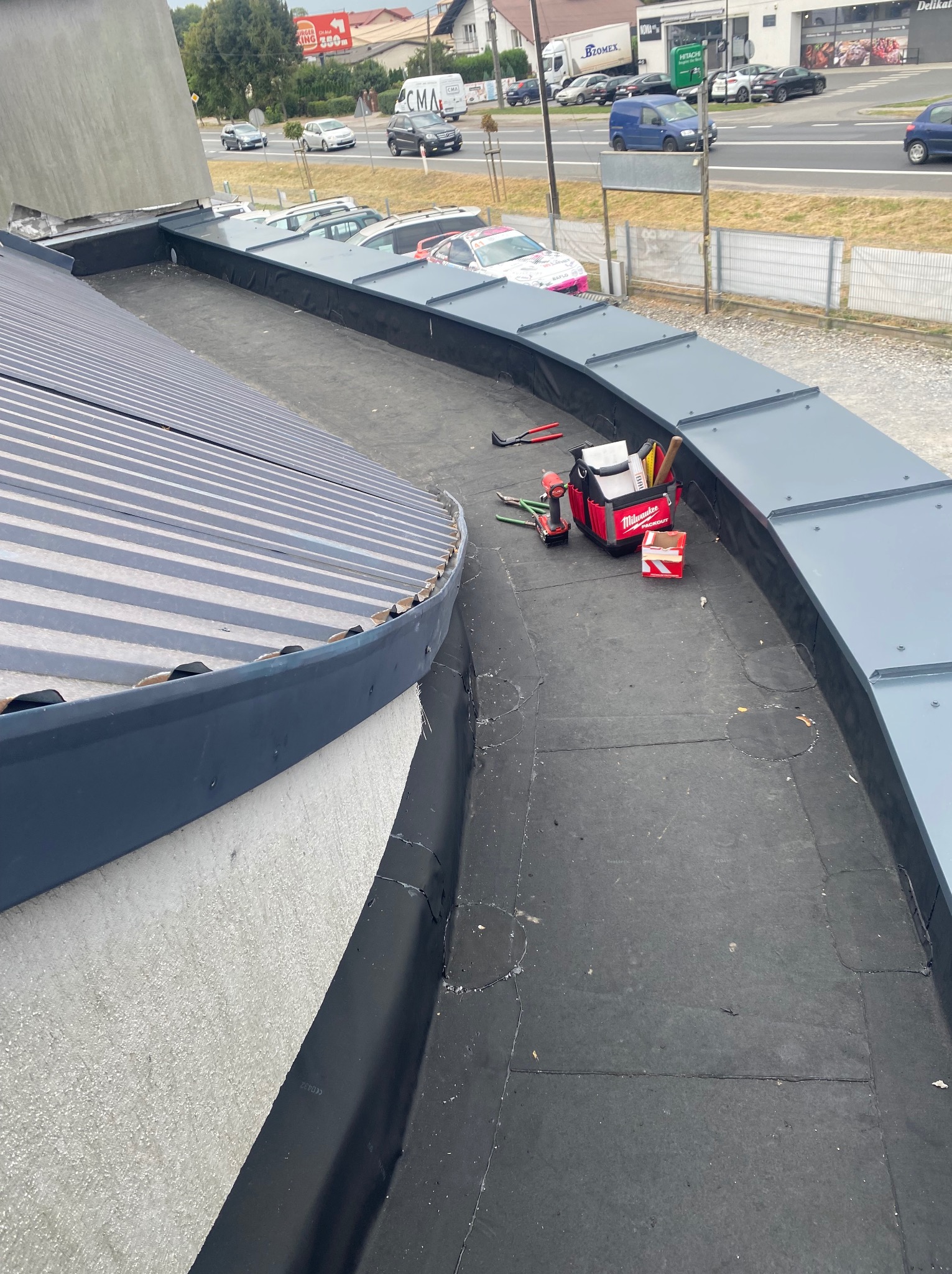 Narzędzia dekarskie, w tym wiertarka akumulatorowa i torba narzędziowa Milwaukee, na płaskim, czarnym dachu z obróbkami blacharskimi w kolorze szarym, widok z góry, w tle zabudowa miejska.