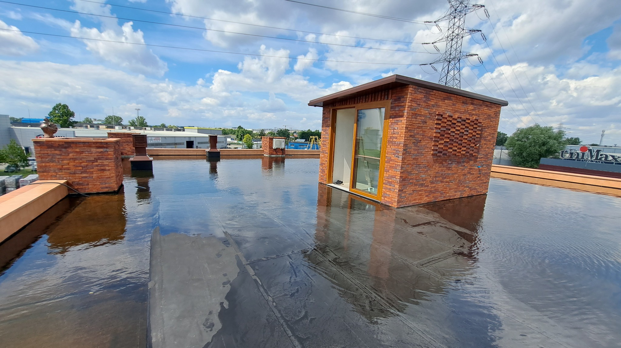 Widok na płaski dach z wykończeniem z cegły klinkierowej, z oknem dachowym i elementami wentylacyjnymi. Na dachu widoczne odbicia nieba i chmur.