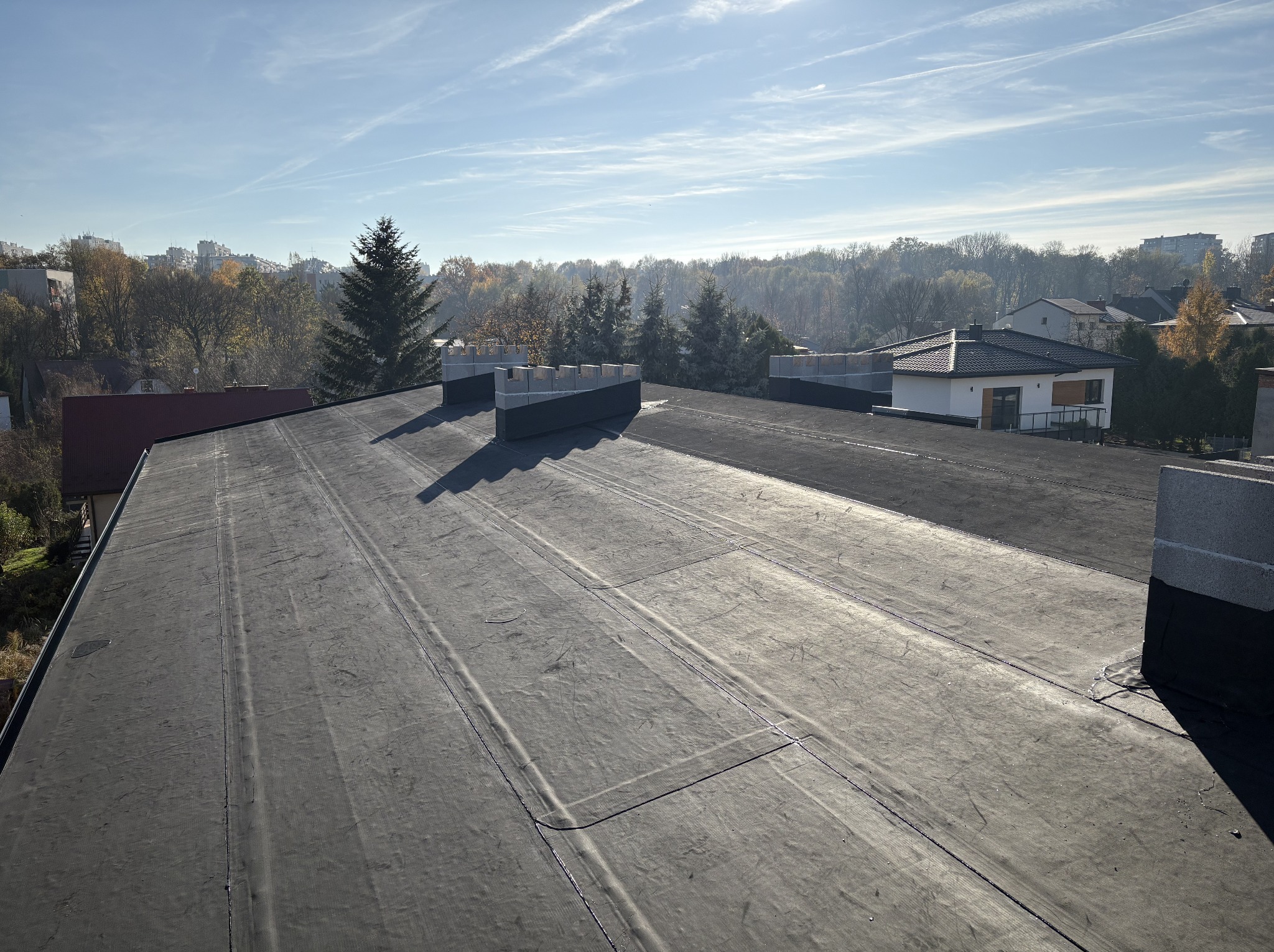 Wykończony, płaski dach pokryty czarną papą termozgrzewalną. Widoczne kominy z szarych bloczków. W tle budynki i drzewa w jesiennej scenerii pod błękitnym niebem.