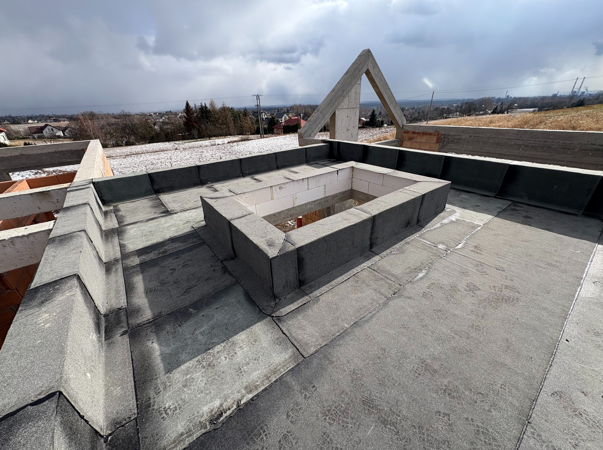 Widok z góry na pokryty papą płaski dach z kominem wentylacyjnym w surowym stanie, w tle krajobraz z zabudowaniami i polem pokrytym śniegiem.