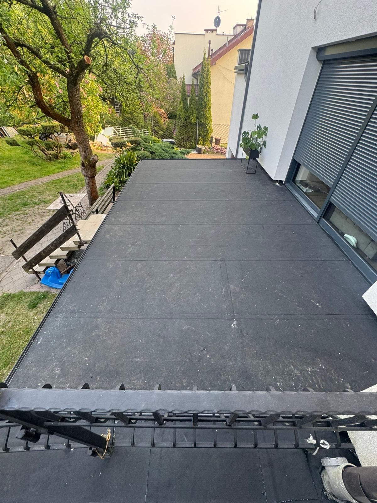 Czarny, płaski dach pokryty papą termozgrzewalną z widocznym ogrodem, elementami architektury i roślinnością w tle, uchwycony z perspektywy balkonu.