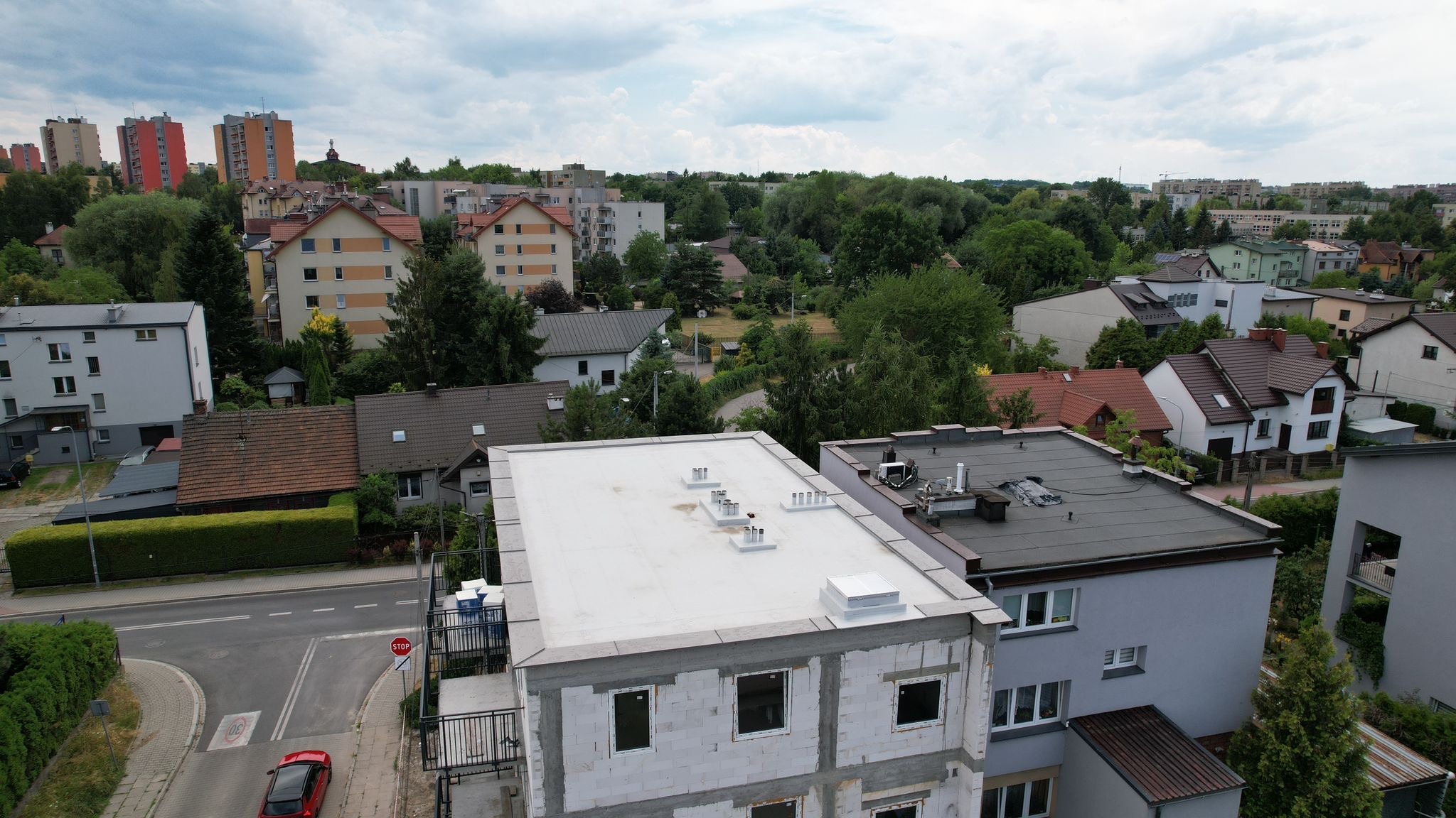 Biały dach płaski z kominkami wentylacyjnymi na budynku w stanie surowym otwartym, widok z góry na tle zabudowy miejskiej i zieleni.