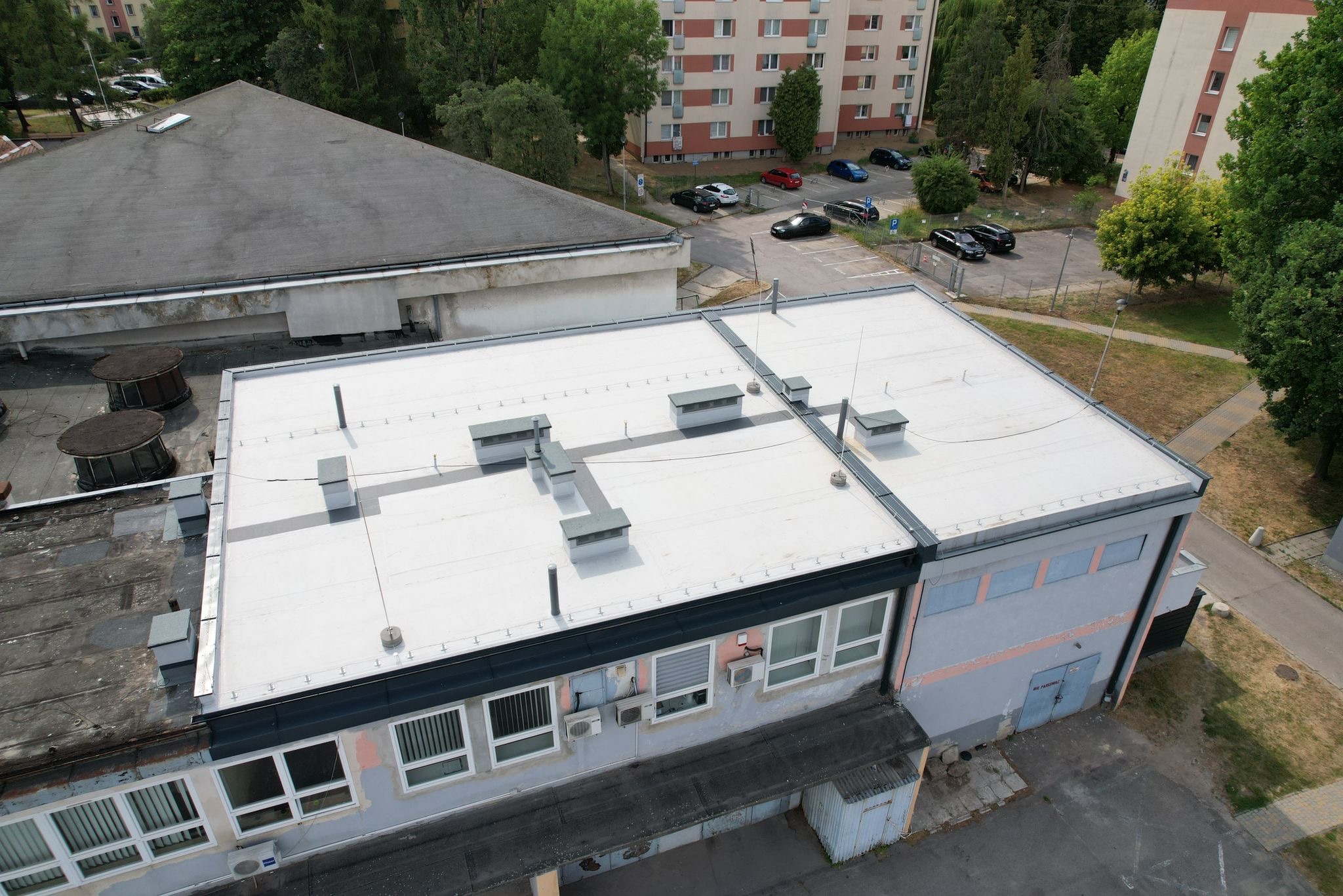 Widok z góry na płaski, biały dach budynku z szarymi elementami wentylacyjnymi i instalacjami. W tle budynki mieszkalne i drzewa. Ujęcie z lotu ptaka.