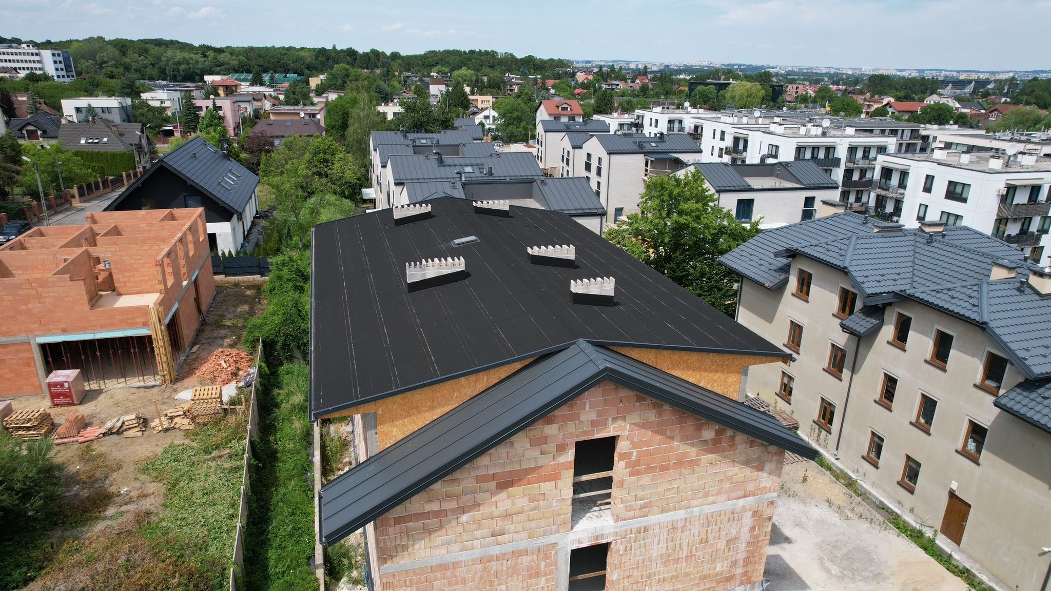 Czarny dach z kominkami wentylacyjnymi na surowym budynku z cegły. Widok z góry na osiedle domów jednorodzinnych w słoneczny dzień.