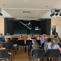 Iluzjonista na scenie prezentuje sztuczkę z długimi, cienkimi rekwizytami przed zgromadzoną widownią dzieci i dorosłych siedzących na krzesłach.