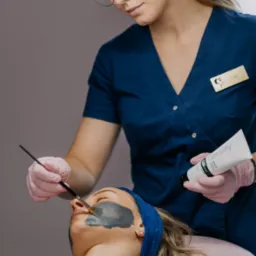 Kosmetyczka w okularach nakłada pędzlem ciemną maskę na twarz klientki leżącej na różowym łóżku kosmetycznym; w dłoni trzyma tubę Detoxify PCA Skin