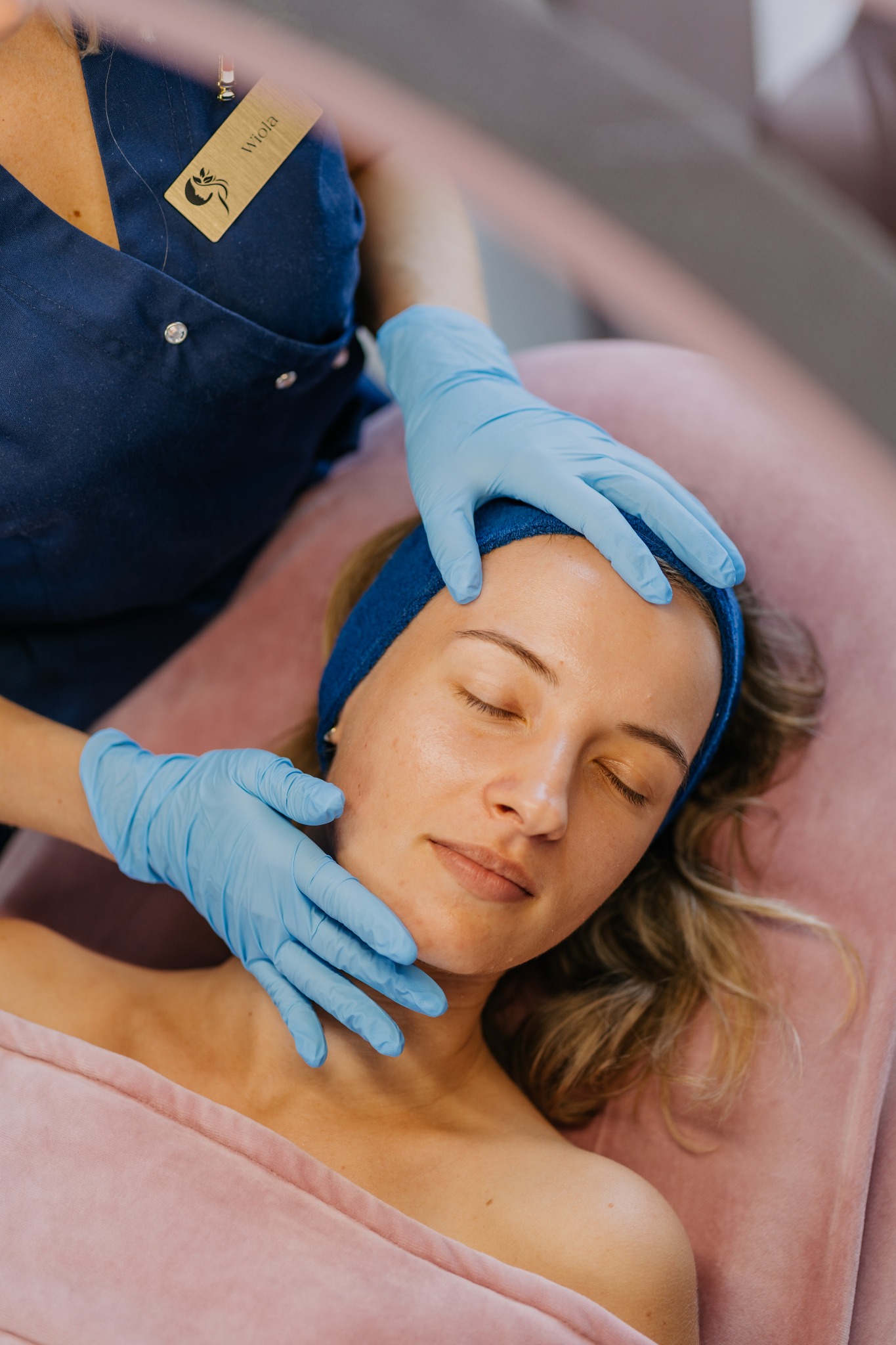 Kobieta z zamkniętymi oczami leży na różowym łóżku, podczas gdy kosmetyczka w niebieskich rękawiczkach wykonuje masaż twarzy. Kosmetyczka ma na sobie niebieski uniform i identyfikator z imieniem...