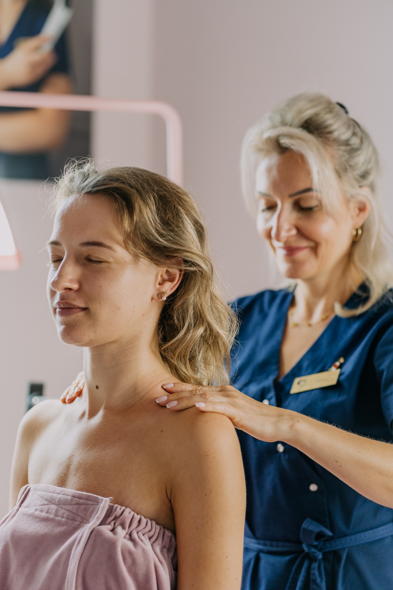 Kobieta w trakcie masażu ramion w gabinecie kosmetycznym; terapeutka w granatowym uniformie z identyfikatorem delikatnie uciska ramiona klientki z zamkniętymi oczami, owiniętej różowym ręcznikiem.