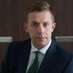 Portret młodego mężczyzny w granatowym garniturze i zielonym krawacie z dyskretnym wzorem, na tle ciemnobrązowej ściany.