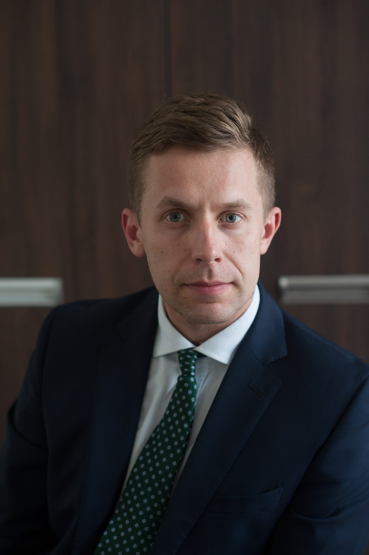 Portret młodego mężczyzny w granatowym garniturze i zielonym krawacie z dyskretnym wzorem, na tle ciemnobrązowej ściany.