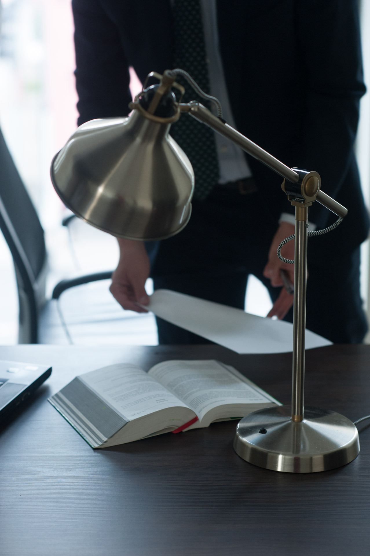 Otwarta księga prawnicza na biurku pod lampką, w tle prawnik w garniturze trzymający dokumenty.