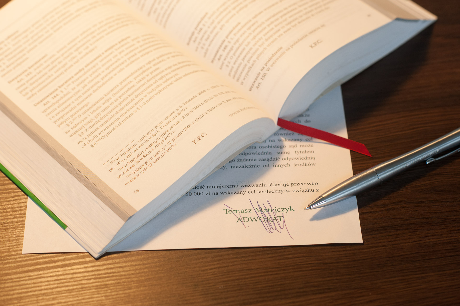 Otwarta książka z aktami prawnymi i srebrny długopis leżące na dokumencie z podpisem adwokata Tomasza Matejczyka na drewnianym biurku.