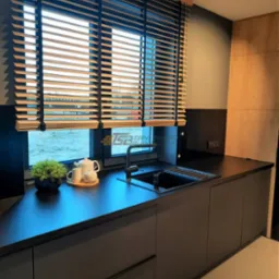 Nowoczesna kuchnia z czarnymi szafkami i blatem, okno z drewnianymi żaluzjami poziomymi, na blacie czajnik i filiżanki.