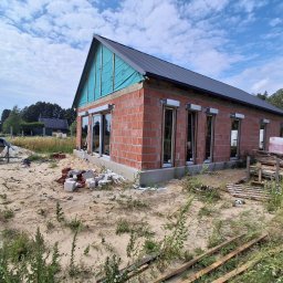 IB_MONSER - Budowa domu z czerwonych pustaków, z zamontowanymi oknami i widocznymi skrzynkami na osłony okienne. Dach z ciemnej blachy, szczyt zabezpieczony niebieską folią.