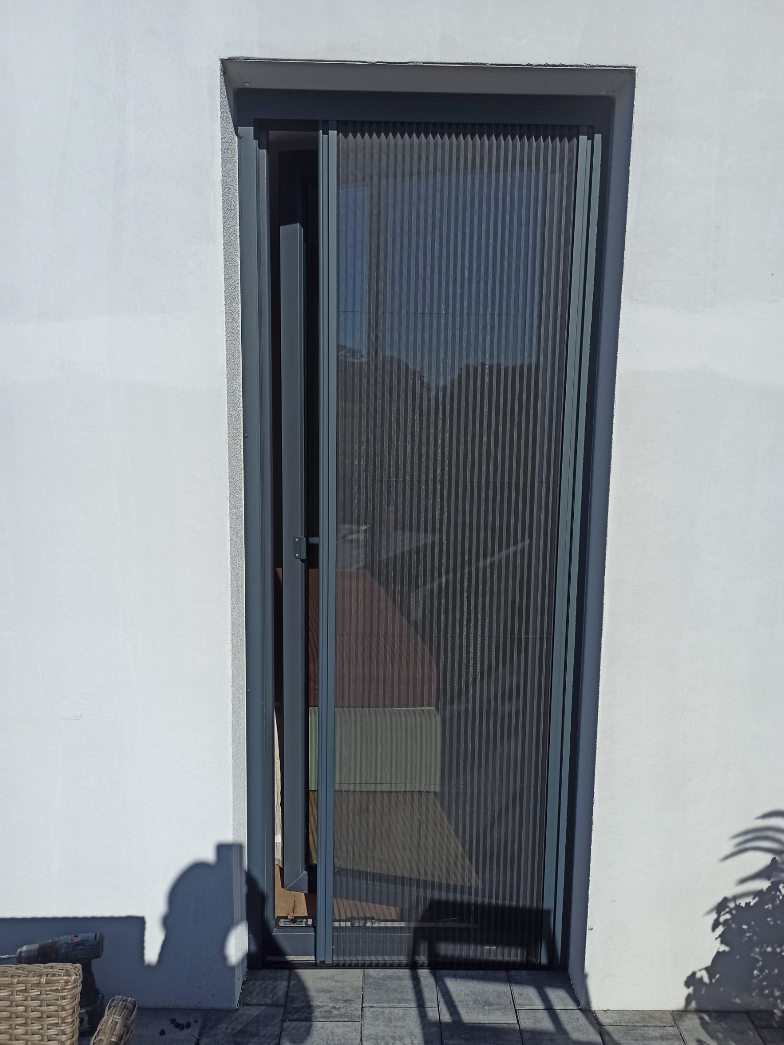 Zamontowana moskitiera plisowana w kolorze grafitowym w Środzie Wielkopolskiej, widoczny fragment ściany budynku i cień osoby.