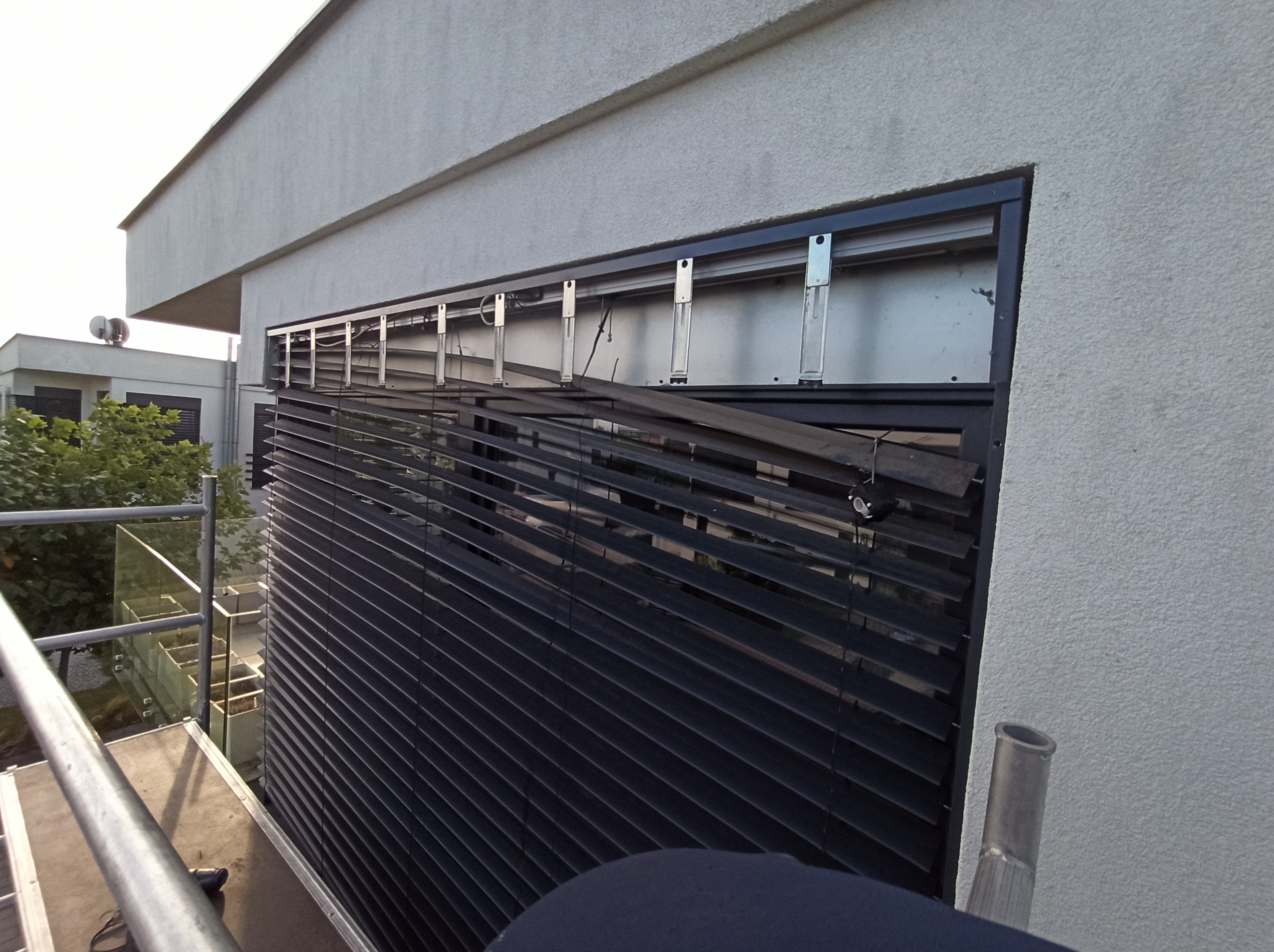 Uszkodzone czarne rolety zewnętrzne zamontowane na elewacji budynku, widoczne elementy konstrukcyjne i częściowo połamane lamele, ujęcie z rusztowania.