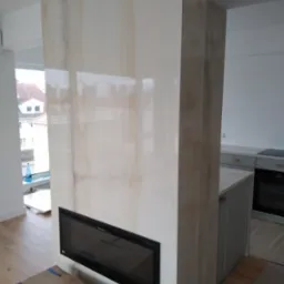 Ściana działowa wykończona płytami imitującymi onyks z wbudowanym biokominkiem, widok z perspektywy podłogi, w tle fragment kuchni i okno.