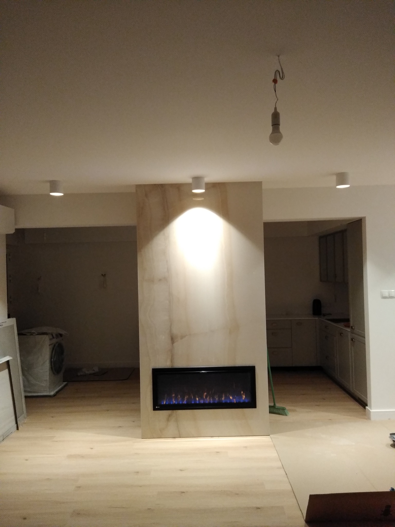 Salon w trakcie wykańczania z elektrycznym kominkiem w obudowie z kamienia, widoczna pralka w tle i niedokończone oświetlenie sufitowe.