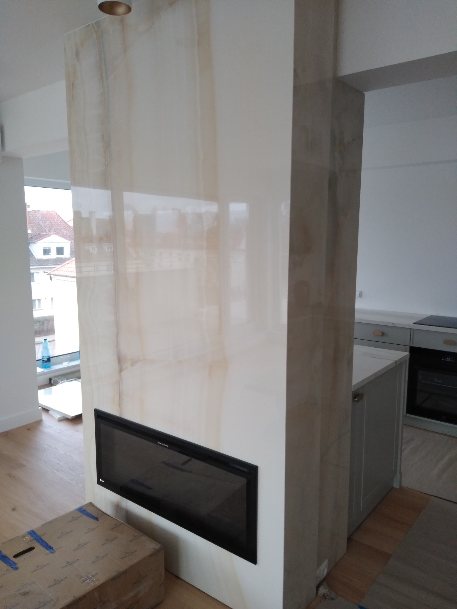 Ściana działowa wykończona płytami imitującymi onyks z wbudowanym biokominkiem, widok z perspektywy podłogi, w tle fragment kuchni i okno.