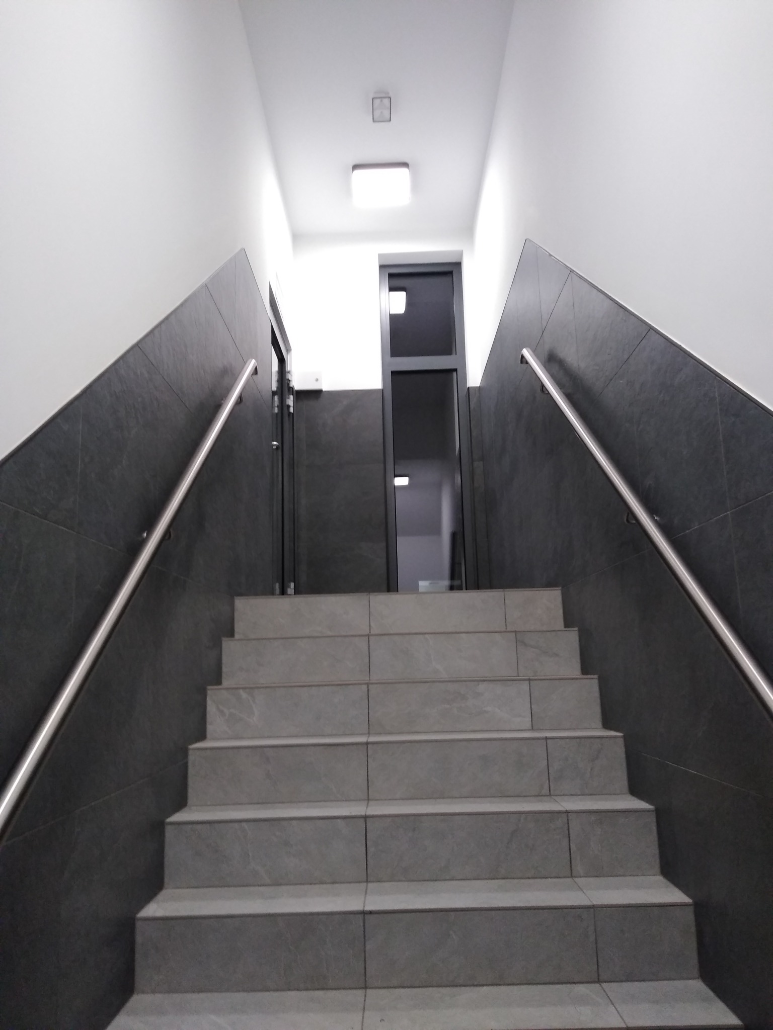 Schody wyłożone szarymi płytkami ceramicznymi, z ciemnoszarymi ścianami i metalową poręczą, prowadzące do drzwi na górze.