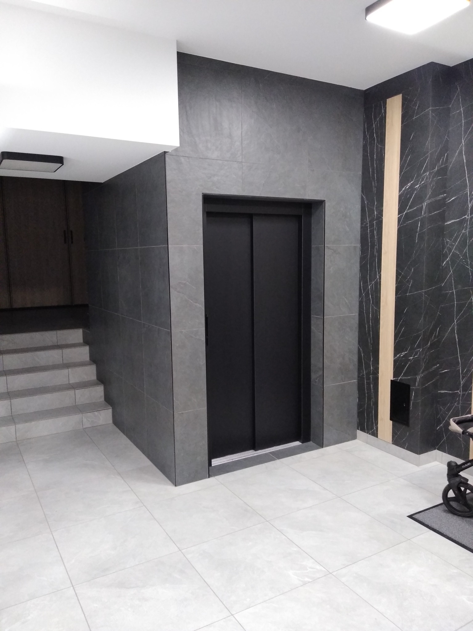 Wykończone płytkami w odcieniach szarości wnętrze z windą, schodami i fragmentem wózka dziecięcego w rogu.