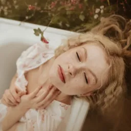 Portret kobiety z blond włosami, leżącej w wannie otoczonej kwiatami, w sukience w drobne kwiaty, z zamkniętymi oczami i rękami skrzyżowanymi na klatce piersiowej, w ciepłej tonacji.