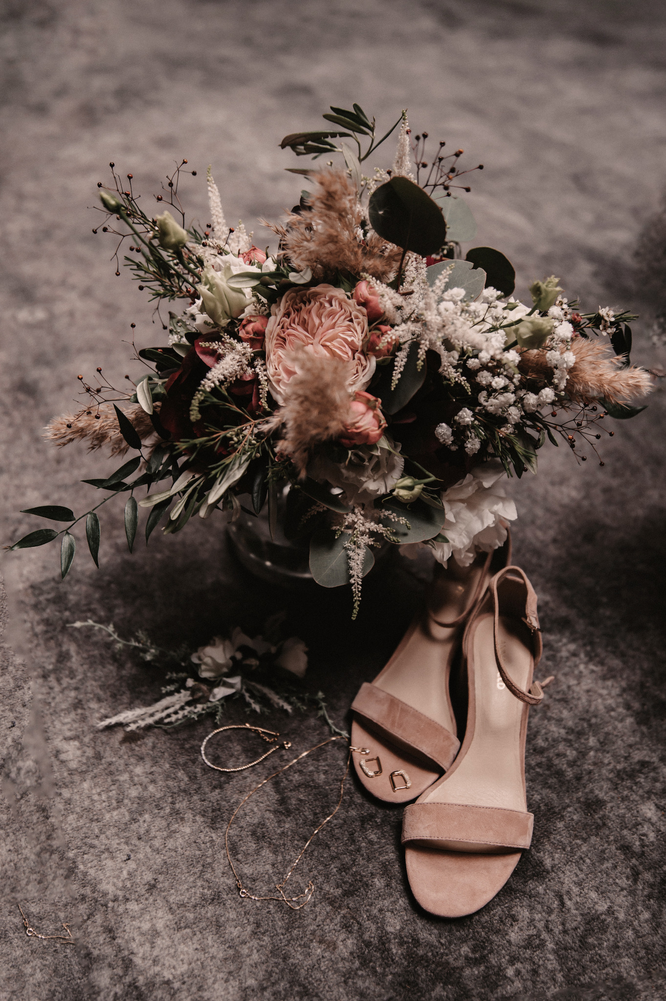 Kompozycja ślubna: bukiet z różowych i białych kwiatów, beżowe sandały na obcasie, złota biżuteria na szarym tle.