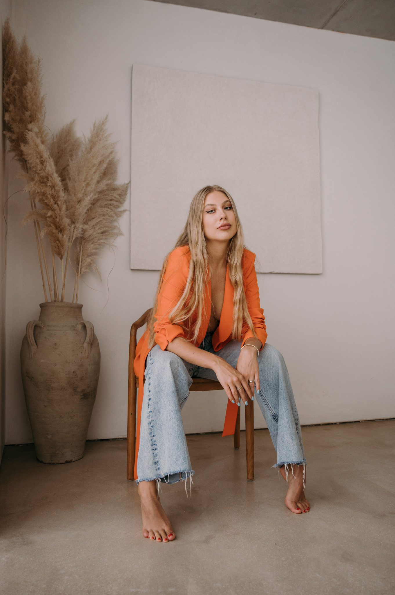 Kobieta z blond włosami siedzi na krześle w pomarańczowej marynarce i jeansach na tle minimalistycznego wnętrza z wazonem z pampasami i białym obrazem.