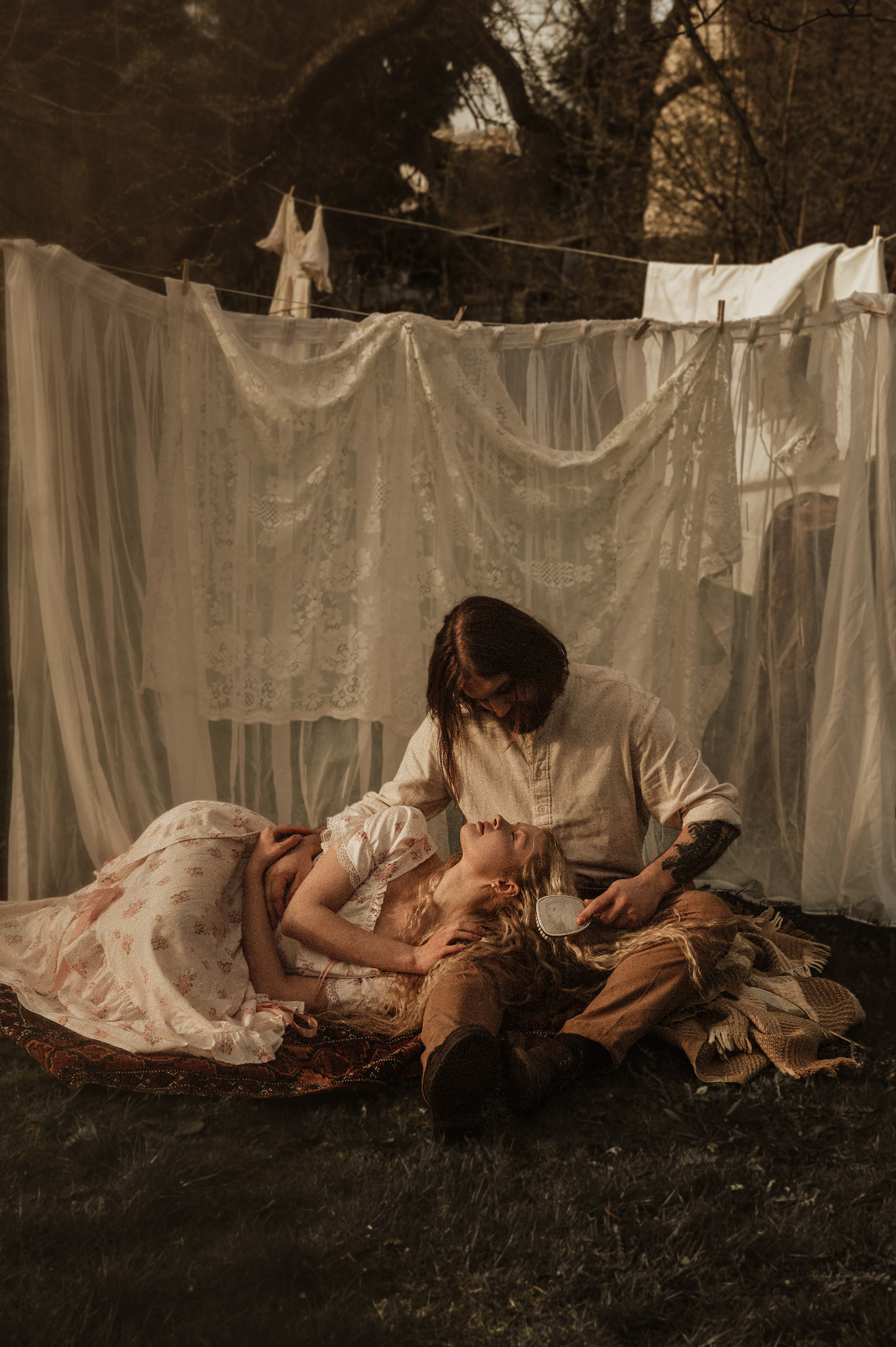 Stylizowana sesja narzeczeńska: mężczyzna czesze włosy leżącej na kocu kobiety na tle rozwieszonego prania z koronkowymi elementami.