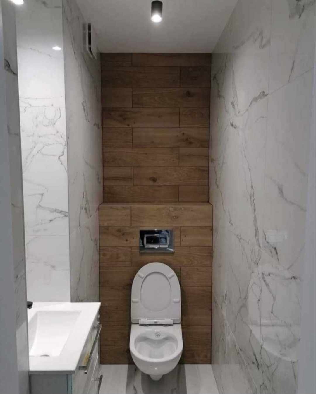 Wnętrze małej łazienki z toaletą podwieszaną na drewnianej ściance, marmurowe płytki na ścianach, umywalka z szafką po lewej stronie, sufit z pojedynczym punktowym oświetleniem.