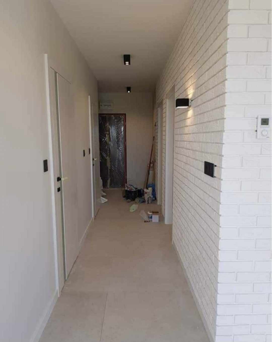 Korytarz w trakcie prac wykończeniowych: biała ceglana ściana z czarnym oświetleniem, gładkie białe ściany, drzwi z czarnymi klamkami, podłoga z jasnych płytek, widoczne narzędzia i materiały...